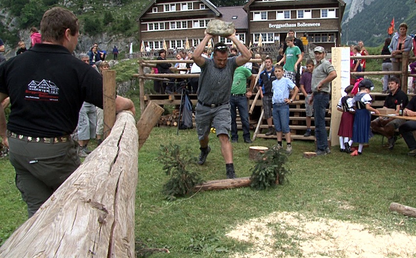 Mann wirft Stein über Kopf dahinter sitzen leute und ein Haus ist sichtbar