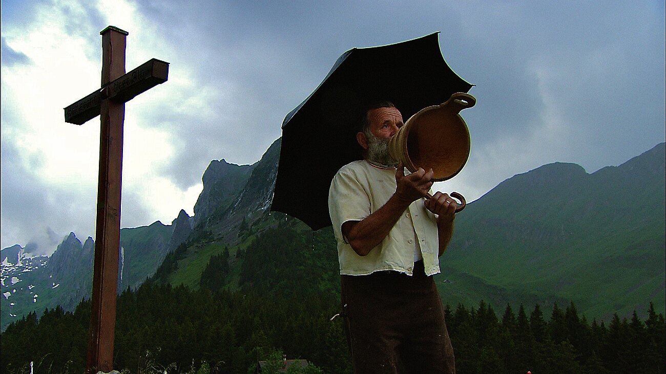 Mann mit Regenschirm singt vor Bergkulisse in ein Holztrichter