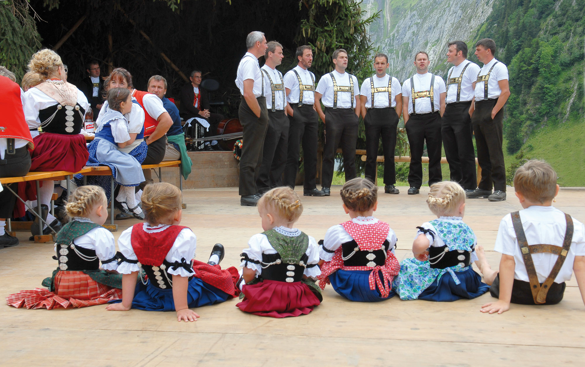 Kinder sitzen vor Gruppe Männer in Tracht und hören ihnen beim Singen zu