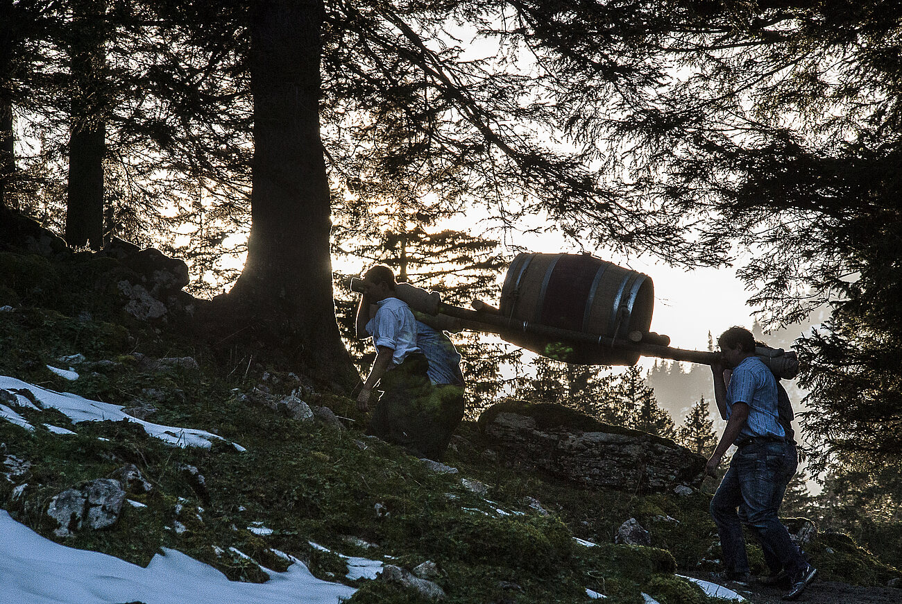 Zwei Männer tragen bei Abendstimmung ein Fass auf den Schultern einen Berg hoch